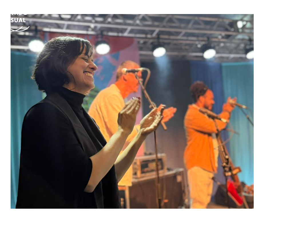 Vânia aparece em primeiro plano na imagem. Ela está em um palco e  a vemos da cintura para cima.


Tem os cabelos curtos na altura da nuca e sorri alegremente. 

Está com as mãos abertas para bater palmas e usa esmalte vermelho.

Ela veste blusa preta de gola alta e blazer preto.

Atrás dela, estão dois músicos, também batendo palmas.