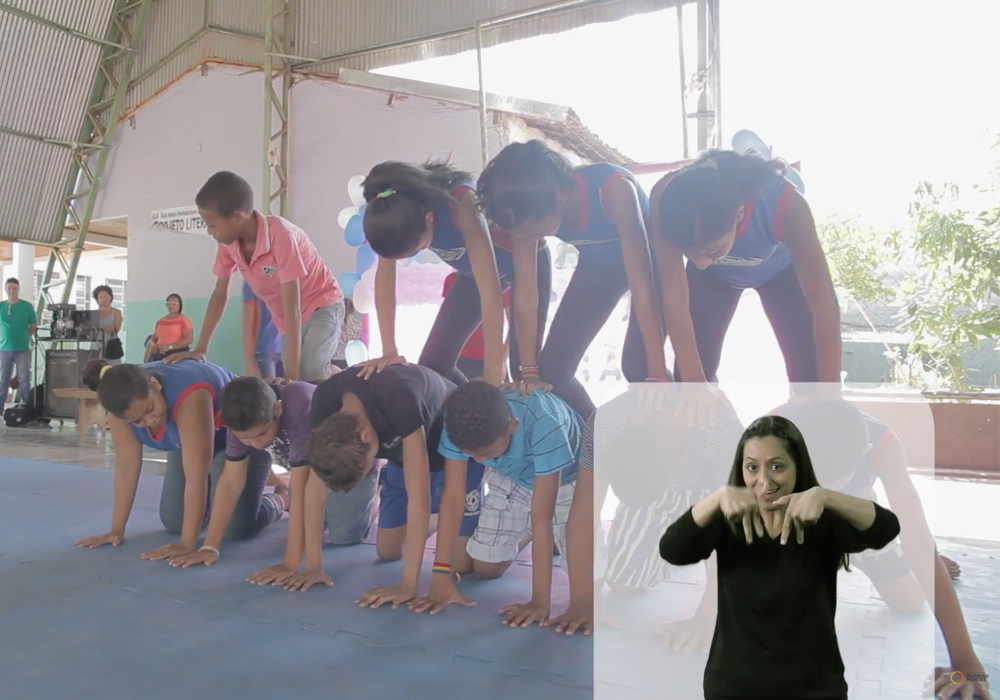 Captura de tela de uma reportagem. 

Nove crianças estão formando uma pirâmide humana, muito comum no circo.

Vânia aparece na Janela de Libras. 

Ela tem os cabelos longos e presos para trás.

Usa uma camiseta preta com mangas longas e calça preta.

Vânia sorri enquanto faz o sinal de [descrever sinal] na Libras.