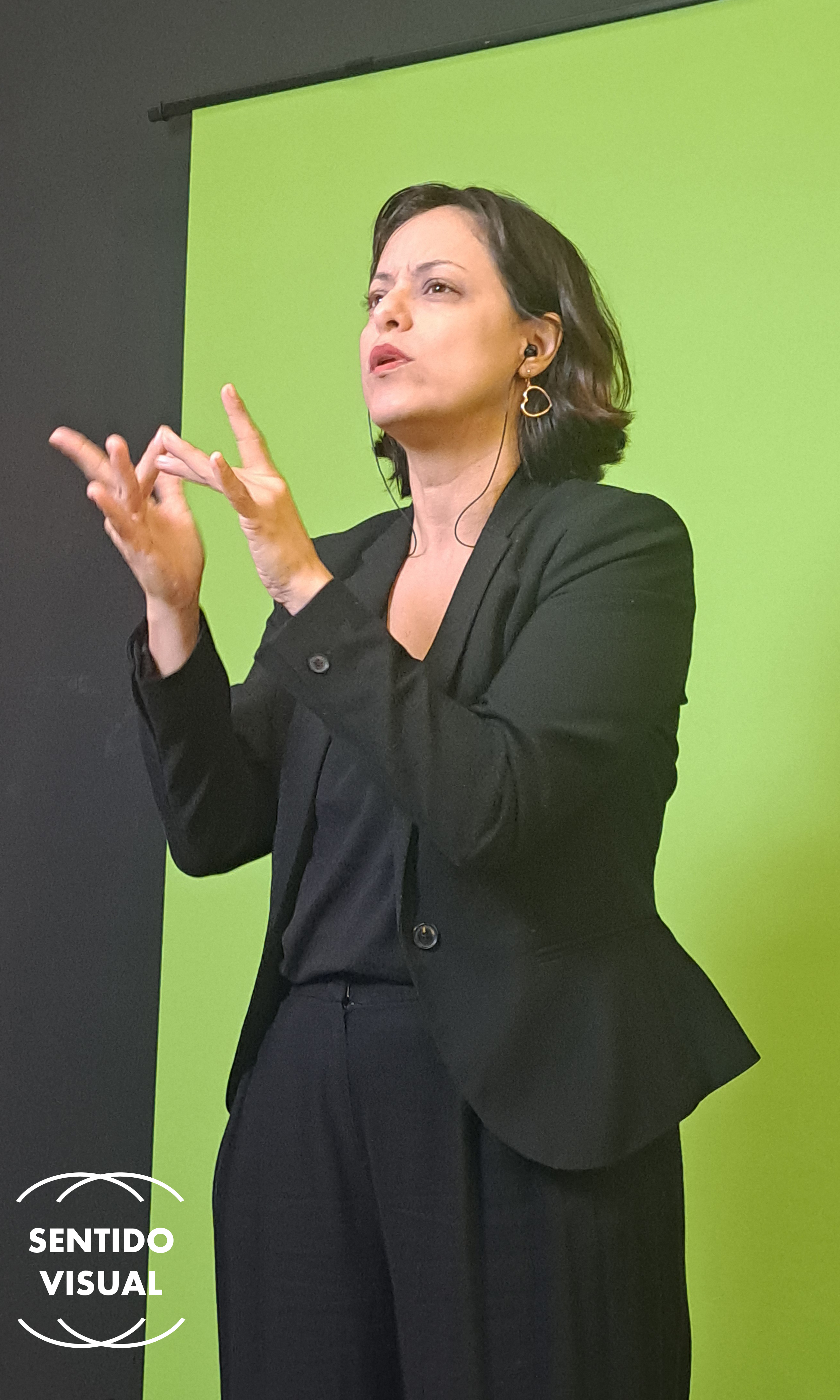 Vânia Santiago em frente a um fundo verde. Ela veste calça social preta, blusa preta e blazer preto. Tem cabelos curtos na altura da orelha e olhos castanhos. Usa um batom nude suave. Na orelha direita, usa um brinco em formato de coração. Ela está fazendo o sinal de contato na Libras: [descrever sinal aqui]